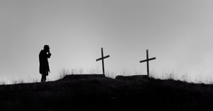 A man mourning on a dreary hill top at the site of two graves marked by crosses.