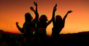 Three women at sunset with hands raised.