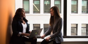 Two women, sitting on a ledge, looking at a laptop.