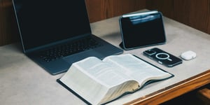 A laptop and a bible open on a wooden desk. 